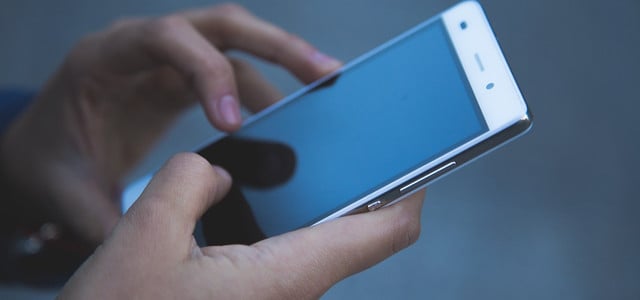 Hände, die ein weißes Smartphone mit schwarzem Bildschirm halten und bedienen, vor unscharfem grauem Hintergrund.