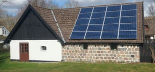 Ein Haus mit einer Fassade aus weißen und steinernen Mauern, braunen Doppeltüren und kleinen Fenstern. Auf dem braunen Ziegeldach sind mehrere blaue Solarmodule installiert.