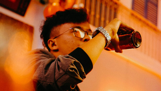 Gen Z trinkt weniger Alkohol als die Generationen vor ihr - das ist der Grund Junger Mann mit Brille und silberner Armbanduhr trinkt aus einer braunen Glasflasche, aufgenommen in warmem, orangefarbenem Licht.