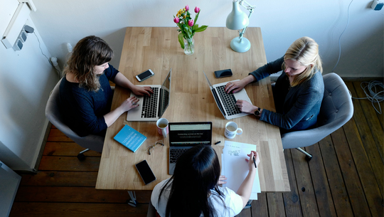 Drei Frauen sitzen an einem Holztisch, zwei arbeiten an Laptops, eine schreibt auf Papier. Auf dem Tisch stehen zwei Kaffeetassen, ein Blumenstrauß, eine Schreibtischlampe, mehrere Smartphones, eine Brille und ein Buch. Der Raum hat Holzboden und weiße Wände.