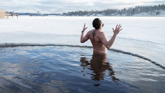 Urlaubstrend: Retreats Ein Mann steht bis zur Taille in einem kreisrunden Loch im zugefrorenen See, umgeben von Eis und Schnee.