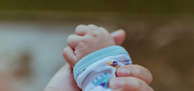 Eine Nahaufnahme einer erwachsenen Hand, die sanft die Hand eines Babys hält. Das Baby trägt ein weißes Kleidungsstück mit einem hellblauen Bündchen und bunten Mustern. Der Hintergrund ist unscharf.