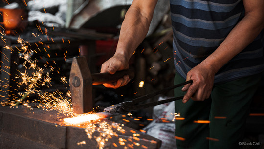 handgeschmiedet Ein Schmied bearbeitet ein glühendes Metallstück mit Hammer und Zange, wobei Funken fliegen. Er trägt ein blau gestreiftes T-Shirt und grüne Hosen.