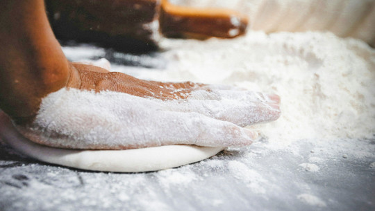 Welche Mehlsorte ist am gesündesten? Eine Hand, die mit Mehl bedeckt ist, drückt einen flachen Teig auf einer bemehlten Arbeitsfläche.