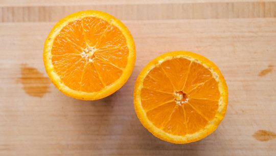 Jeden Tag Orangen: Das sind die Folgen Zwei halbierte Orangen liegen mit der Schnittfläche nach oben auf einem hellen Holzbrett.