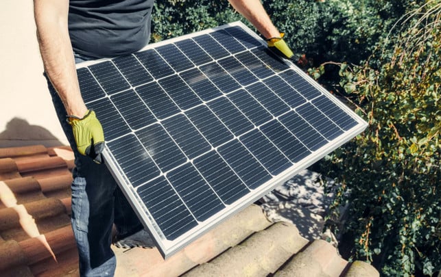 251013-groesse-gewicht-anzahl-solarzellen-cc0-pexels-kindel-media Person mit gelben Arbeitshandschuhen hält ein Solarpanel auf einem Dach mit roten Dachziegeln.