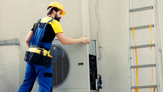 Heizungsgesetz: Wie geht es weiter? Ein Handwerker mit gelbem Schutzhelm, gelb-schwarzem T-Shirt und blauer Arbeitslatzhose öffnet das Gehäuse einer Wärmepumpe an einer Außenwand.