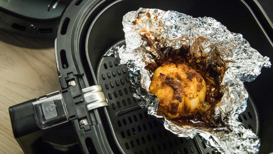 Teils brandgefährlich: 8 Airfryer-Fehler und wie du sie vermeidest Ganze Knoblauchknolle, gebacken in Alufolie, in einem Heißluftfritteusen-Korb. Die Knolle ist leicht gebräunt und karamellisiert.