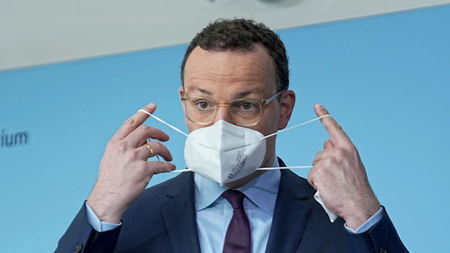 Spahn-maskendeals-dpa Ein Mann mit Brille, Anzug und Krawatte zieht eine weiße FFP2-Maske über sein Gesicht. Er hält die Gummibänder der Maske mit beiden Händen an den Ohren. Der Hintergrund ist hellblau.