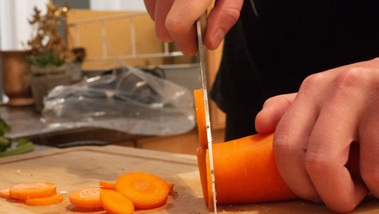 Jeden Tag Karotten essen: Das sind die Folgen Eine Hand schneidet mit einem Messer eine Karotte in dünne Scheiben auf einem Holzbrett. Mehrere Karottenscheiben liegen bereits auf dem Brett. Im Hintergrund sind unscharf Küchenutensilien und Pflanzen zu erkennen.