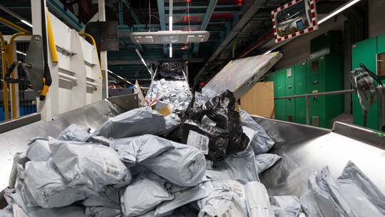 EU will 2026 Abgabe auf Billig-Importe einführen: wichtiger Schritt gegen Temu und Co. Internationales Postzentrum (IPZ) am Frankfurter Flughafen: Förderband mit zahlreichen verpackten Paketen in grauen und schwarzen Kunststoffhüllen in einer Lager- oder Sortieranlage, im Hintergrund grüne Schaltschränke und eine Überwachungsspiegel an der Wand.