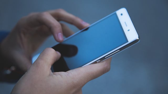 Hände, die ein weißes Smartphone mit schwarzem Bildschirm halten und bedienen, vor unscharfem grauem Hintergrund.