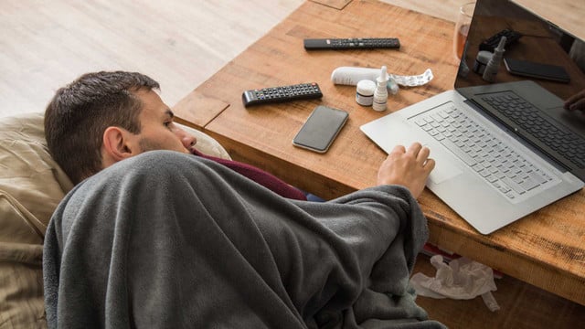 Online-Attest kann zu Kündigung führen: Vermeide diesen Fehler Ein Mann liegt mit einer grauen Decke zugedeckt auf einem Sofa und benutzt einen silbernen Laptop auf einem Holztisch. Auf dem Tisch liegen zwei Fernbedienungen, ein Smartphone, mehrere Medikamentenbehälter und eine zerknüllte Tablettenverpackung. Unter dem Tisch sind gebrauchte Papiertaschentücher sichtbar.