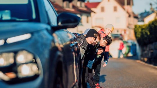 Schulwege-Index 2025 Fünf Kinder sehen hinter einem blauen Auto hervor, das auf einer sonnigen Straße geparkt ist. Die Kinder tragen bunte Jacken und Mützen.
