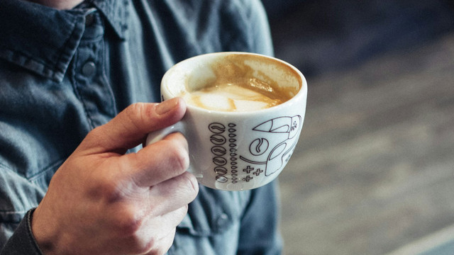 Die beliebteste Kaffeesorte der Welt ist in Gefahr Hand, die eine weiße Tasse mit Kaffeeresten und Milchschaum hält, die Tasse ist mit schwarzen Kaffeebohnen- und Symbolzeichnungen verziert, im Hintergrund ein dunkelblaues Hemd.