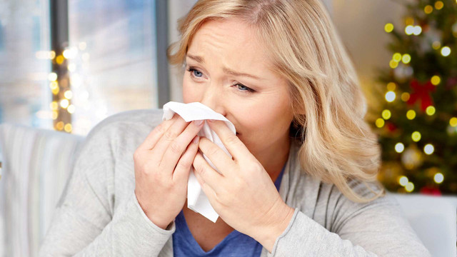 Krank an Weihnachten: Was tun, wenn die Arztpraxis geschlossen ist? Frau mit blonden Haaren, die sich mit einem Taschentuch die Nase putzt, im Hintergrund unscharfer Weihnachtsbaum mit Lichtern.