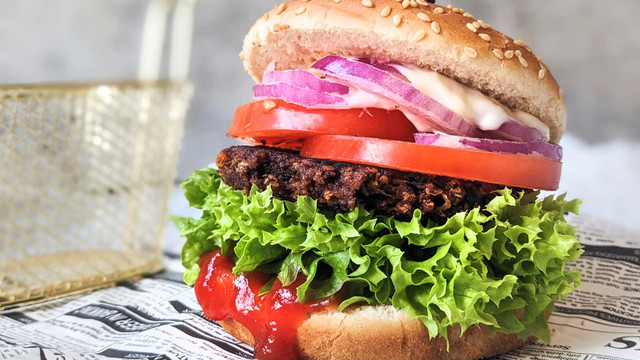 EU vertagt Entscheidung: Bleibt der Veggie-Burger ein Burger? Hamburger mit veganem Patty und Sesambrötchen, grünem Salat, Tomatenscheiben, roten Zwiebeln, einer dunklen, knusprigen Pattieschicht, Mayonnaise und Ketchup, serviert auf Zeitungspapier.