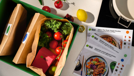 HelloFresh_1920x1080_2 Eine grüne Box mit drei braunen Papiertüten, von denen eine geöffnet ist und frisches Gemüse wie Tomaten, Brokkoli und Kräuter sowie eine rote HelloFresh-Verpackung enthält. Daneben liegen drei Rezeptkarten mit den Titeln "Yellow Thai Style Veggie Noodles", "Mediterranean Salmon" und "Japanese umami glazed chicken". Auf der Arbeitsfläche sind außerdem eine Zitrone, eine rote Zwiebel, eine kleine Dose und weitere Tomaten zu sehen.
