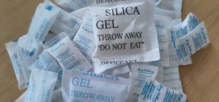 Mehrere kleine Beutel mit Silicagel, beschriftet mit Warnhinweisen wie "DESICCANT", "SILICA GEL" und "DO NOT EAT", liegen auf einer Holzoberfläche.
