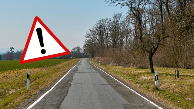 Vorsicht mit dem Auto: Diese Tiere sind jetzt wieder vermehrt auf den Straßen unterwegs Eine schmale Landstraße mit unebener, reparierter Asphaltoberfläche, umgeben von Wiesen und kahlen Bäumen. Links im Bild ist ein großes, rotes Warndreieck mit einem schwarzen Ausrufezeichen zu sehen.