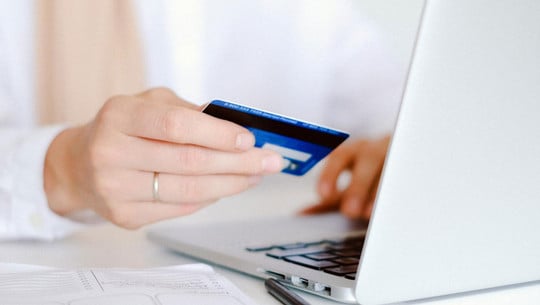 Zum 16. Mal in Folge: Das ist die "Bank des Jahres" 2025 Hand mit silbernem Ring hält eine blaue Kreditkarte vor einem geöffneten silbernen Laptop, auf einem weißen Tisch liegen ein Stift und ein Notizbuch.