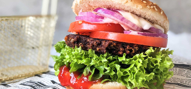 Hamburger mit veganem Patty und Sesambrötchen, grünem Salat, Tomatenscheiben, roten Zwiebeln, einer dunklen, knusprigen Pattieschicht, Mayonnaise und Ketchup, serviert auf Zeitungspapier.