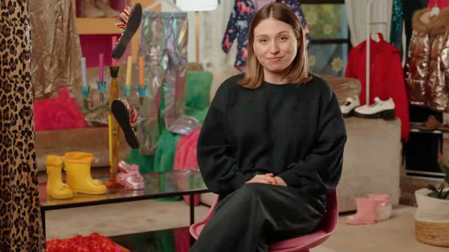 Früher ließ sie sich einen LKW voll Shein-Kleidung liefern – jetzt rechnet "Bibi" mit Ultra Fast Fashion ab Bianca Heinicke sitzt auf einem pinken Stuhl. Sie trägt einen schwarzen Pullover und einen schwarzen Rock. Im Hintergrund sind bunte Kleidungsstücke, darunter ein Leopardenmuster, gelbe Gummistiefel, ein roter Mantel, weiße Schuhe und pinke Stiefel zu sehen. Außerdem sind ein Kerzenhalter mit bunten Kerzen und ein Tisch mit roten Tomaten darauf zu erkennen.