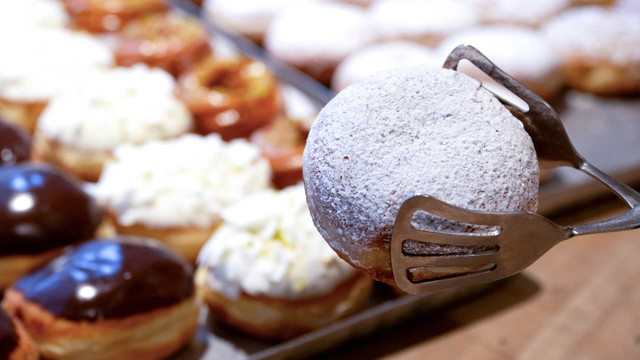 Vorsicht vor dieser Krapfenfüllung: Amt kritisiert Hygiene in 11 von 18 Proben Ein mit Puderzucker bestäubter Berliner (Krapfen) wird mit einer Metallzange gehalten, im Hintergrund sind weitere Berliner mit verschiedenen Glasuren und Toppings unscharf zu sehen.