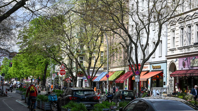 Studie zu Kiezblocks: Diese positiven Wirkungen hat Verkehrsberuhigung Die Bergmannstraße im Bergmannkiez in Berlin-Kreuzberg. Belebte Straße mit mehreren geparkten Autos und Radfahrern, umgeben von Gebäuden mit bunten Markisen. Bäume mit teilweise belaubten Ästen säumen den Straßenrand, und es sind verschiedene Verkehrsschilder, darunter ein Stoppschild, sichtbar. Auf der rechten Seite sind Cafés oder Geschäfte mit Außenbereichen und Sitzgelegenheiten zu sehen.
