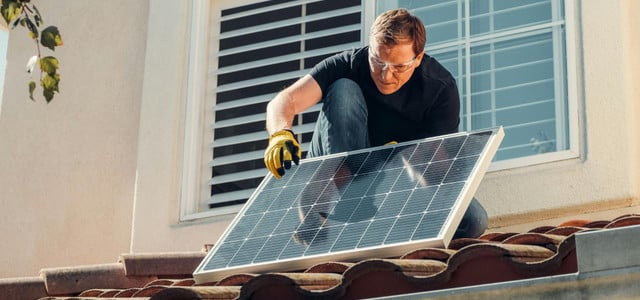 Ein weißer Mann mit Schutzbrille und Arbeitshandschuhen installiert ein Solarpanel auf einem Dach mit roten Ziegeln .