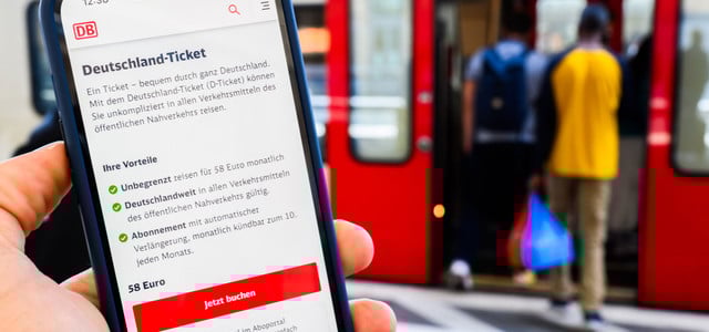 Hand, die ein Smartphone mit der Buchungsseite für das Deutschland-Ticket der Deutschen Bahn zeigt, im Hintergrund steigen zwei Personen in eine rote Bahn ein.