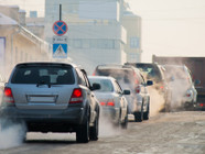 autos-abgase-verbrenner-cb-yury-260120 Autos und ein Lastwagen auf einer Straße, aus den Auspuffen sind Abgase sichtbar. Im Hintergrund sind Gebäude und ein Verkehrsschild.