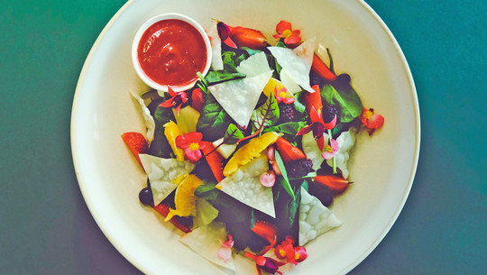 Weniger Fleisch essen: brauchst du nahrungsergänzungsmittel? Bunter Salat mit Spinat, Erdbeeren, Orangenstücken, essbaren Blüten und knusprigen Dreiecken, serviert mit einer kleinen Schale roter Sauce auf einem weißen Teller.