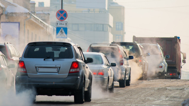 autos-abgase-verbrenner-cb-yury-260120 Autos und ein Lastwagen auf einer Straße, aus den Auspuffen sind Abgase sichtbar. Im Hintergrund sind Gebäude und ein Verkehrsschild.