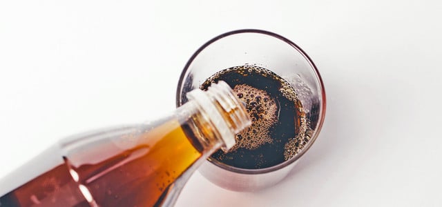 Eine braune Flasche gießt eine dunkle, kohlensäurehaltige Flüssigkeit in ein durchsichtiges Glas auf weißem Hintergrund.