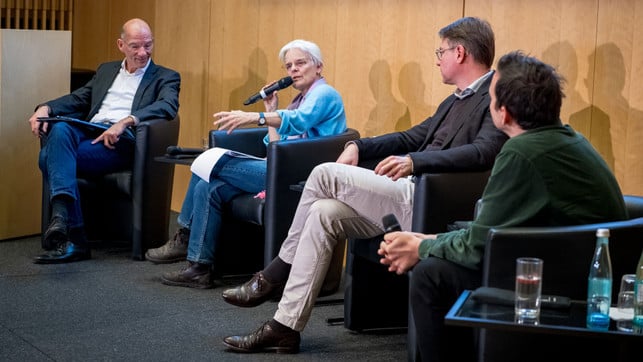 FairFinanceWeek_OSC2735_1920x1080 Vier Personen sitzen in Sesseln auf einer Bühne vor einer Holzpaneelwand. Eine ältere Frau mit weißem Haar und blauer Strickjacke spricht in ein Mikrofon und gestikuliert mit der Hand. Die anderen drei Personen hören ihr aufmerksam zu. Auf einem kleinen Tisch neben ihnen stehen Wasserflaschen und Gläser.