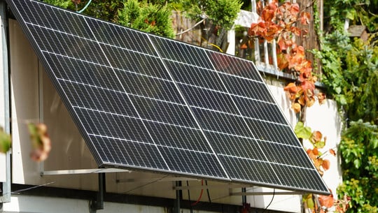 251020-speicher-balkonkraftwerk-sinnvoll-c Zwei Solarpaneele sind an ein Balkongeländer montiert, umgeben von grünen und herbstlich gefärbten Pflanzen.