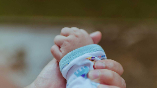 „Was stimmt nicht mit mir?“: Wieso viele ihre Elternschaft bereuen – und was hilft Eine Nahaufnahme einer erwachsenen Hand, die sanft die Hand eines Babys hält. Das Baby trägt ein weißes Kleidungsstück mit einem hellblauen Bündchen und bunten Mustern. Der Hintergrund ist unscharf.