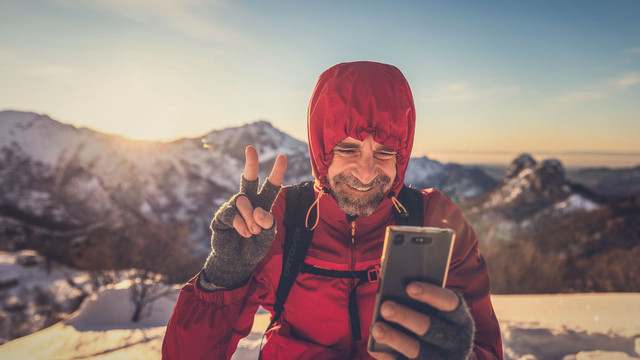 1920x1080_Mann_in-_den_Bergen_web Ein Mann in roter Winterjacke mit Kapuze und grauen Handschuhen hält ein Smartphone in der Hand und zeigt mit der anderen Hand ein Peace-Zeichen. Er steht in einer verschneiten Berglandschaft bei Sonnenuntergang.