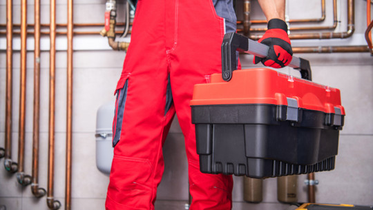 Heizungsgesetz: Wichtige Regel ab 2026 Heizungsmonteur in roter Arbeitskleidung hält einen schwarzen und roten Werkzeugkasten vor einer Wand mit Heizungsrohren