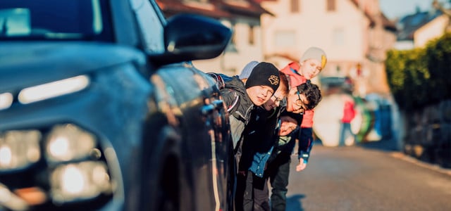 Fünf Kinder sehen hinter einem blauen Auto hervor, das auf einer sonnigen Straße geparkt ist. Die Kinder tragen bunte Jacken und Mützen.