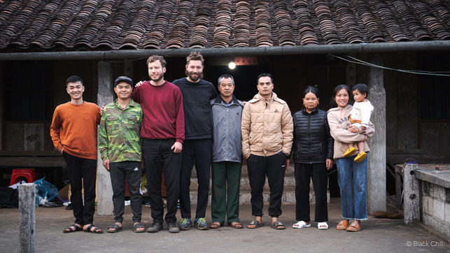 black-chili-familie-vietnam Eine Gruppe von neun Personen steht vor einem traditionellen Gebäude mit einem Ziegeldach. Die Personen tragen verschiedene Freizeitkleidung, darunter Pullover, Jacken und Sandalen. Eine Frau hält ein kleines Kind im Arm. Alle blicken in die Kamera und einige lächeln.