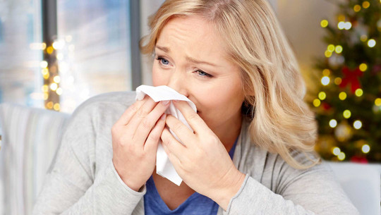 Krank an Weihnachten: Was tun, wenn die Arztpraxis geschlossen ist? Frau mit blonden Haaren, die sich mit einem Taschentuch die Nase putzt, im Hintergrund unscharfer Weihnachtsbaum mit Lichtern.