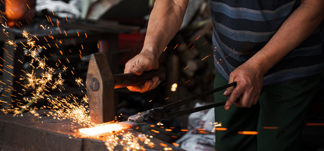 Circular Economy: Wie alter Stahl in deiner Küche weiterlebt. ((© Black Chili)) Ein Schmied bearbeitet ein glühendes Metallstück mit Hammer und Zange, wobei Funken fliegen. Er trägt ein blau gestreiftes T-Shirt und grüne Hosen.