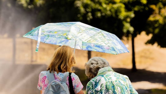 Bericht: Millionen Menschen sterben wegen Klimakrise – eine Maßnahme rettet etwa 160.000 Leben pro Jahr Zwei Frauen von hinten, eine mit roten Haaren und Rucksack, die andere mit grauen Haaren, die gemeinsam einen bunten Regenschirm mit floralen Mustern halten. Im Hintergrund unscharfe Bäume und ein sonniger Tag.