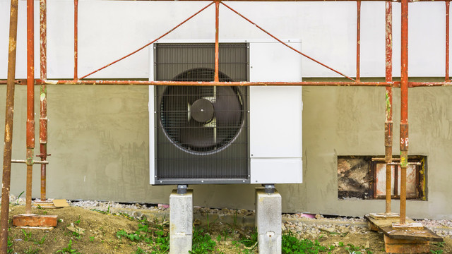 Mit Wärmepumpe und energetischer Sanierung sparen Außeneinheit einer Wärmepumpe, montiert auf zwei Betonblöcken vor einer Hauswand, umgeben von Baugerüst