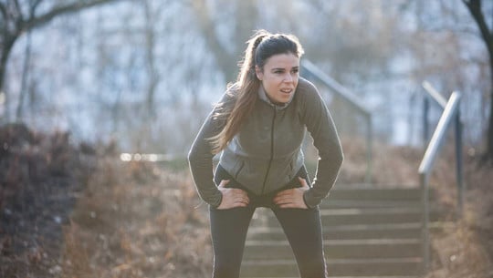 Schmerzen bei Sport? Experten verraten, was hilft - und was du besser nicht tun solltest Junge Frau in Sportkleidung lehnt sich mit den Händen auf den Knien nach vorne, wirkt erschöpft, steht vor einer Treppe im Freien an einem bewölkten Tag.