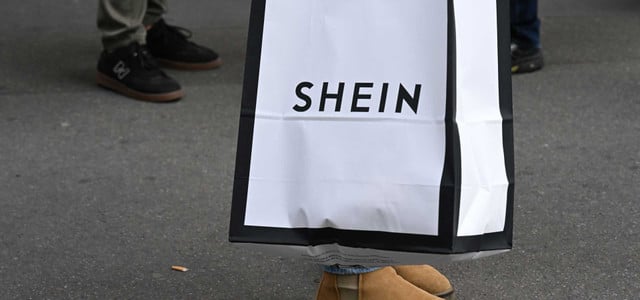 Frankreich weist Shein in die Schranken – wie reagiert Deutschland? (Foto: Julien Mattia/Le Pictorium via ZUMA Press/dpa) Eine Einkaufstüte mit dem Aufdruck "SHEIN" in schwarzer Schrift auf weißem Hintergrund mit schwarzem Rand, gehalten von einer Person mit braunen Wildlederstiefeln auf grauem Asphalt. Im Hintergrund sind weitere Personen mit dunklen Schuhen zu sehen.