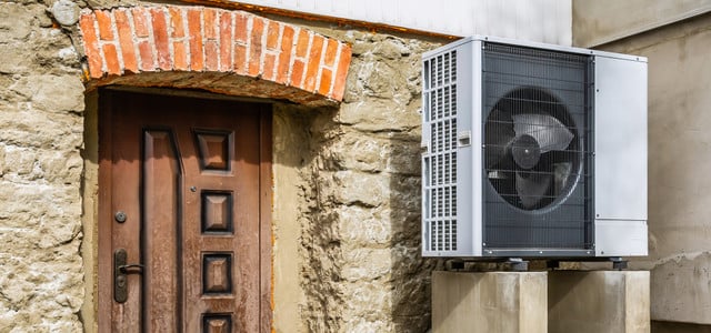Langzeitstudie: Wärmepumpen sind auch im Altbau effizient (Foto: © Colourbox.de) Außeneinheit einer Wärmepumpe, montiert auf zwei Betonblöcken neben einer alten Steinmauer mit einer braunen Holztür und einem Bogen aus roten Ziegelsteinen darüber.