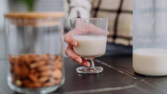 BfR warnt vor Schimmelpilzgiften: Für wen Mandelmilch gefährlich sein kann Hand hält ein Glas mit pflanzlicher Milch, daneben steht ein Glasbehälter mit Mandeln und ein weiterer Behälter mit pflanzlicher Milch auf einem dunklen Tisch.
